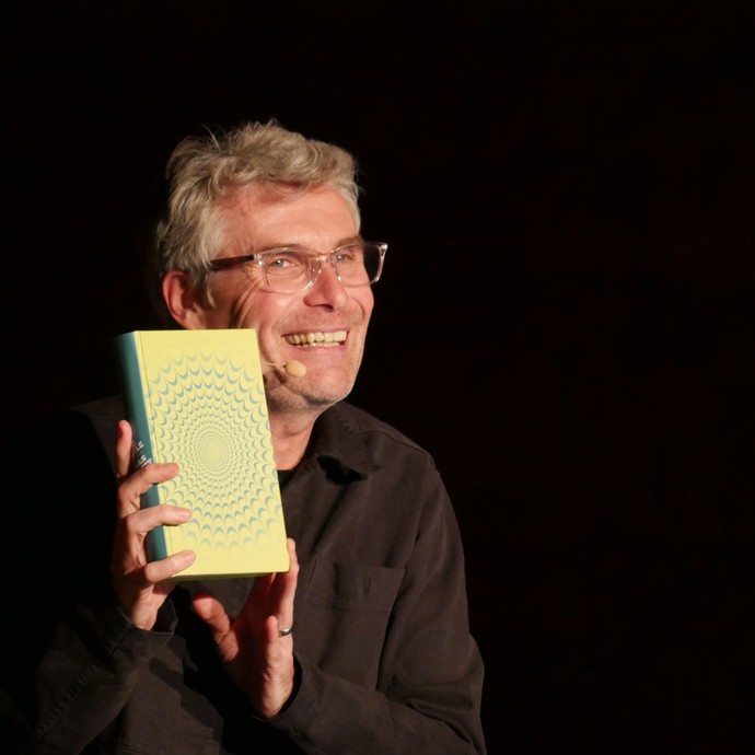 Der Schauspieler Harald Schwaiger in seiner Rolle als Achtsamkeitstrainer hält lachend ein Buch hoch. (vergrößerte Bildansicht wird geöffnet)