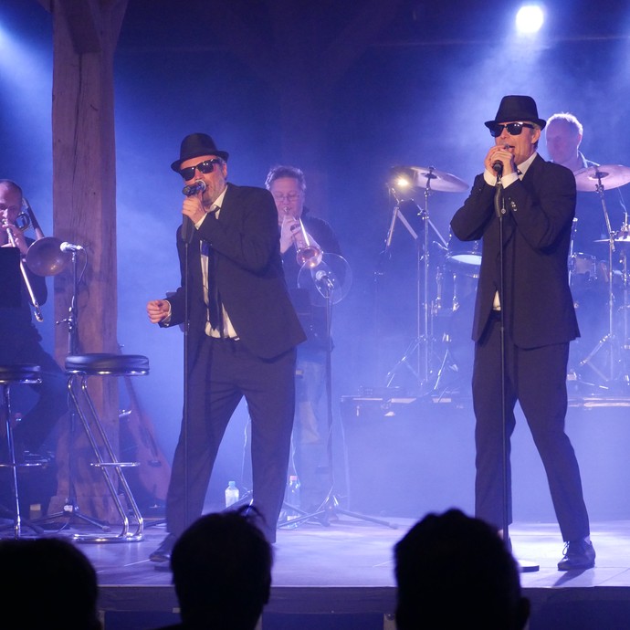 Die Schauspieler Michael Kamp und Harald Schwaiger singen als Blues Brothers vor einer Band. (vergrößerte Bildansicht wird geöffnet)