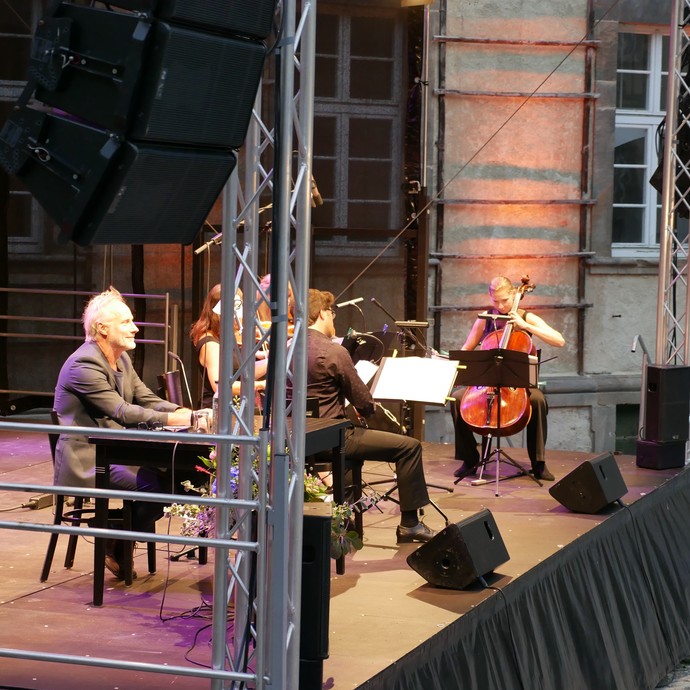 Der Schauspieler Wolfram Koch sitzt an einem Tisch auf der Bühne. Neben ihm spielen mehrere Instrumentalisten. (öffnet vergrößerte Bildansicht)